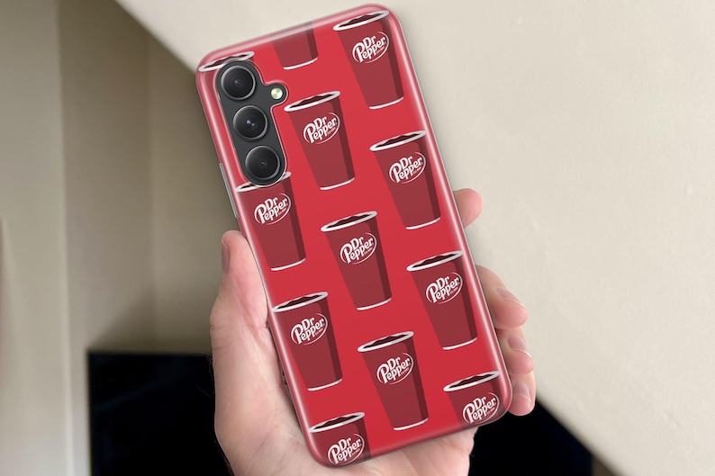Puede incluir: Funda de tel&eacute;fono roja con un patr&oacute;n repetido de vasos de refresco Dr. Pepper. Los vasos son de color rojo oscuro con texto blanco y un borde blanco. La funda tiene una c&aacute;mara de triple lente.