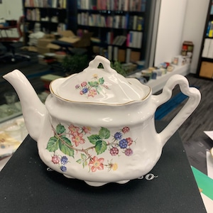 May include: White teapot with a floral design featuring pink, blue, and green flowers and blackberries. The teapot has a gold trim around the lid and spout.