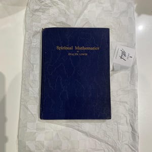 Puede incluir: Un libro azul marino titulado "Spiritual Mathematics" de Evalyn Lowes. El libro está sobre una superficie blanca y texturizada. Una pequeña etiqueta blanca con texto negro es visible a la derecha del libro.