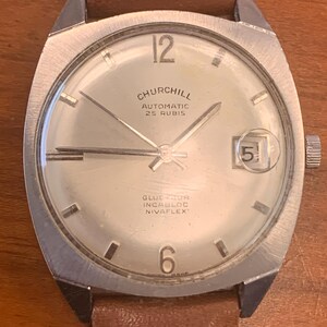 May include: A silver-toned wristwatch with a brown leather strap. The watch face is white with black markings and the text "Churchill Automatic 25 Rubis Glucydur Incabloc Nivarflex".