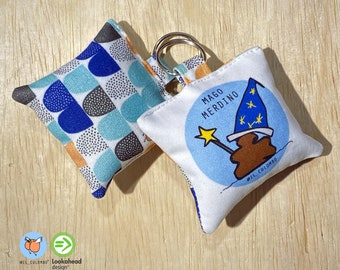 Pode incluir: Um chaveiro de tecido branco com um círculo azul e o texto "Mago Merdino" em preto. O círculo apresenta uma ilustração de desenho animado de uma figura marrom usando um chapéu azul com estrelas e segurando uma varinha mágica. O chaveiro está preso a um chaveiro de tecido branco menor com um padrão geométrico azul, cinza e laranja.