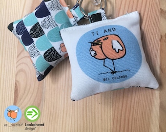 Pode incluir: Um chaveiro de tecido branco e azul com um desenho de pássaro de desenho animado. O pássaro é laranja e tem o texto "TI ANO" acima dele. O chaveiro também tem um quadrado de tecido menor com um padrão geométrico em tons de azul, verde e preto.
