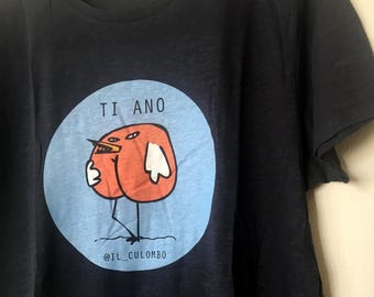 Pode incluir: T-shirt azul marinho com um desenho de pássaro de desenho animado dentro de um círculo azul claro. O pássaro é laranja com detalhes brancos e pernas pretas. O texto "TI ANO" está acima do pássaro e "@IL_CULOMBO" abaixo.