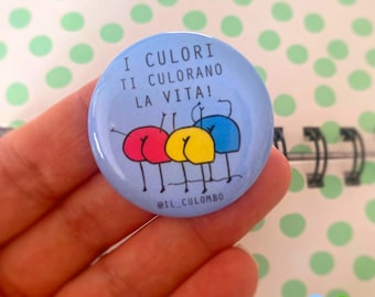 Pode incluir: Um botão azul claro com o texto "I COLORI TI COLORANO LA VITA!" e uma ilustração de desenho animado de três traseiros coloridos. Os traseiros são vermelhos, amarelos e azuis. O botão é segurado em uma mão.
