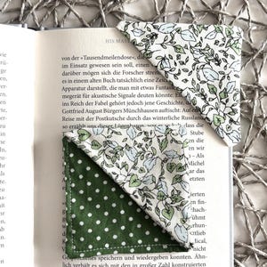 Op de afbeelding: Twee handgemaakte stoffen bladwijzers. Eén is groen met witte stippen, de andere is wit met een groen bloemenpatroon. De bladwijzers zijn in een boek geplaatst met zichtbare tekst. De achtergrond is een geweven mat.