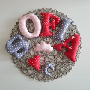 May include: A handmade fabric banner with the letters "SOFIA" in pink and red, with a cloud, heart, and star shapes in grey and red. The banner is hanging on a woven rope.