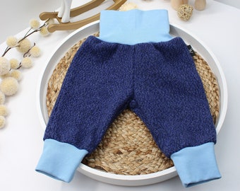 May include: Navy blue baby pants with light blue cuffs and waistband. The pants are made of a textured fabric and are displayed on a white tray with a woven base. Decorative items include a white and gold rocking horse and mushroom figurines.
