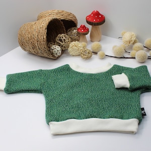 May include: A green textured sweater with white trim on the cuffs, neckline, and hem. The sweater is displayed on a white surface, with decorative elements including a woven basket, wooden mushrooms, and floral accents in the background.