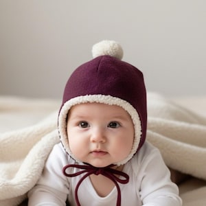 May include: A deep burgundy bonnet with a cream-coloured sherpa lining and a matching pom-pom on top. The bonnet features thin burgundy ties that fasten under the chin, providing a snug fit around the face while keeping the head warm and covered.