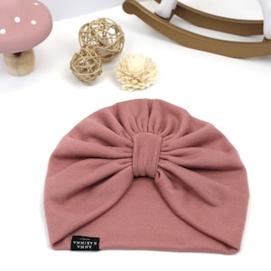 May include: A dusty rose turban-style baby hat with a large bow detail. The hat is made of soft fabric and features a small black tag with white text that reads "ANNA KARINNA". The hat is designed for infants and toddlers.