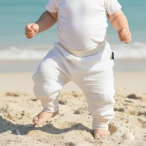 May include: A baby wearing a light blue textured bucket hat, a plain white short-sleeved bodysuit, and off-white muslin cotton pants with a gathered waistband and cuffed ankles, standing on a sandy beach with the ocean in the background.