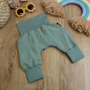 May include: Sage green baby trousers with a gathered, textured fabric. The trousers have a wide waistband and cuffs at the ankles. The trousers are made of a soft material and are designed for comfort. The trousers are laying on a wooden surface.