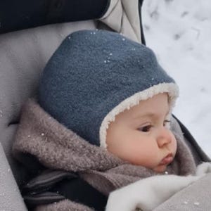 Peut inclure: Un enfant est assis dans une poussette gris foncé, portant un bonnet en laine bleue avec une doublure en polaire blanche et une veste en polaire texturée marron. La poussette présente une doublure intérieure gris clair et une chancelière assortie.