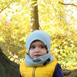 Peut inclure: Un enfant porte un bonnet bleu clair et un tour de cou assorti, ainsi qu'un gilet jaune. L'arrière-plan présente un arbre et des feuilles jaunes, suggérant une scène d'automne. Le bonnet et le tour de cou ont de petites étiquettes noires.