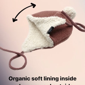 Peut inclure: Un bonnet en laine marron avec un pompon blanc, des oreillettes et de longs liens. L'int&eacute;rieur pr&eacute;sente une doublure en polaire blanche. Une &eacute;tiquette noire avec le texte ANNA KARINNA est visible. Le texte Organic soft lining inside and warm wool outside est pr&eacute;sent.