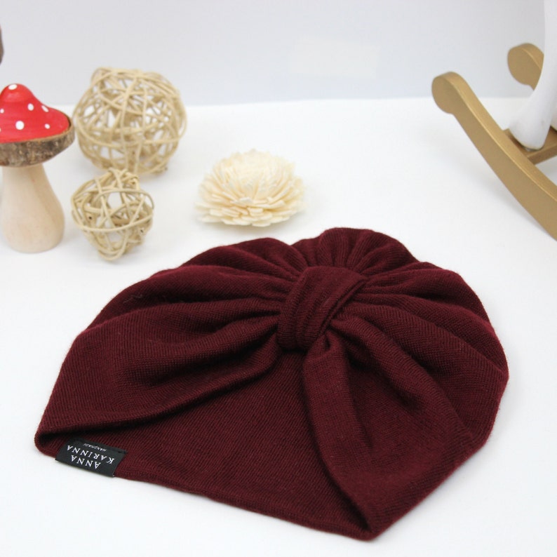 May include: A burgundy-colored turban-style hat with a knotted bow detail. The hat has a small black tag with white text that reads "ANNA KARINA". The hat is made of a soft, knitted material.