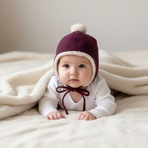 May include: A deep burgundy bonnet with a cream-coloured sherpa lining and a matching pom-pom on top. The bonnet features thin burgundy ties that fasten under the chin, providing a snug fit around the face while keeping the head warm and covered.
