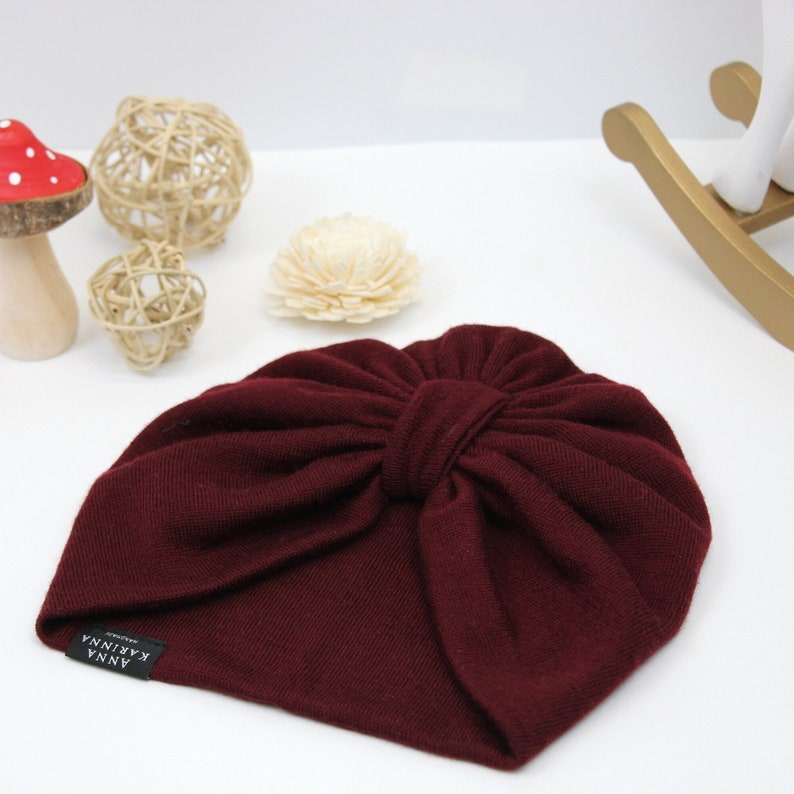 May include: A burgundy-colored baby turban with a knotted bow detail. The hat has a soft, knitted texture and a small black label with white text that reads "ANNA KARINNA". The turban is a stylish accessory for infants.