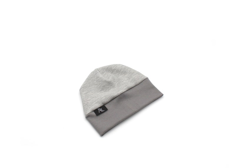 Może przedstawiać: Szara czapka beanie z żebrowanym wierzchem i gładkim szarym rondem. Czapka ma małą czarną metkę z białym napisem. Czapka wykonana jest z miękkiego, dzianinowego materiału i nadaje się na zimną pogodę.