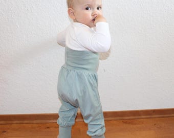 May include: A baby wearing a white long-sleeved shirt and light blue pants. The pants have a high waist and gathered ankles. The baby is standing on a wooden floor.