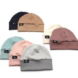 Peut inclure: Collection de sept bonnets de différentes couleurs : marine, beige, vert d'eau, rose, crème, mauve et gris. Chaque bonnet présente une texture côtelée et une petite étiquette noire avec du texte blanc. Les bonnets sont présentés sur fond blanc.