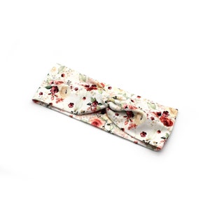 May include: A floral headband featuring a cream background with a pattern of red, pink, and green flowers and leaves. The headband is knotted at the top, creating a stylish accessory.