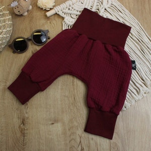 May include: Burgundy-colored baby pants with a high waistband and ribbed cuffs. The pants are made of a textured fabric and are displayed on a wooden surface. A small tag with the letters "AK" is visible.