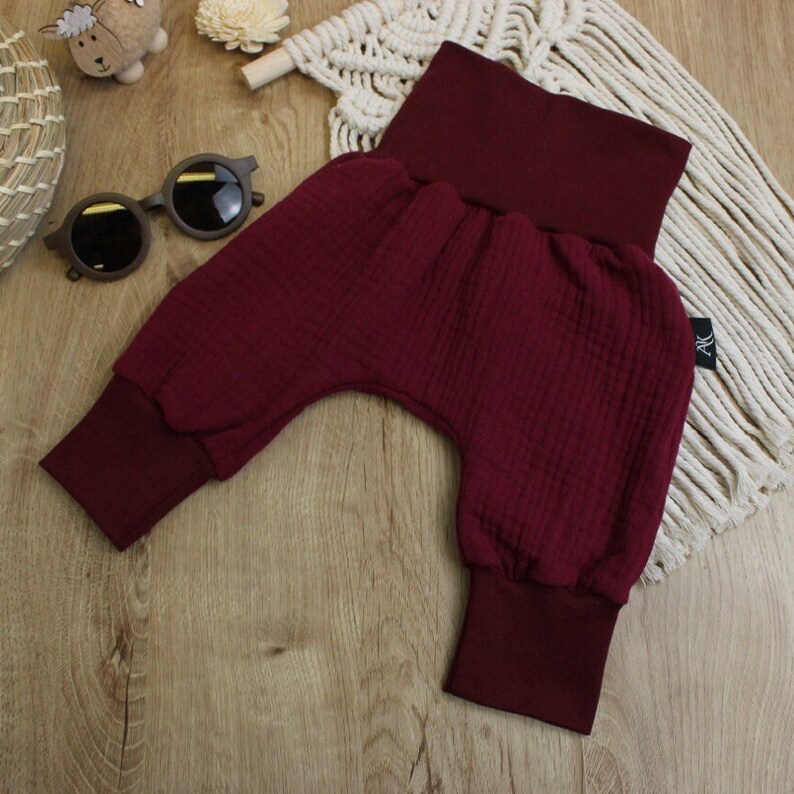 May include: Burgundy-colored baby pants with a high waistband and ribbed cuffs. The pants are made of a textured fabric and are displayed on a wooden surface. A small tag with the letters "AK" is visible.