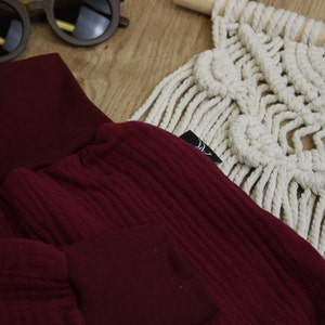 May include: Close-up of a burgundy ribbed knit fabric, possibly clothing, with a matching solid fabric detail. Wooden sunglasses and a cream-colored macrame wall hanging are in the background. A black tag is attached to the fabric.