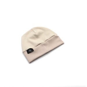 Może przedstawiać: Prążkowana czapka beanie w dwukolorowym designie. G&oacute;rna część jest w kolorze jasnobeżowym, a wywinięty brzeg w ciemniejszym, stonowanym beżu. Mała czarna metka z białym tekstem jest wszyta na brzegu.