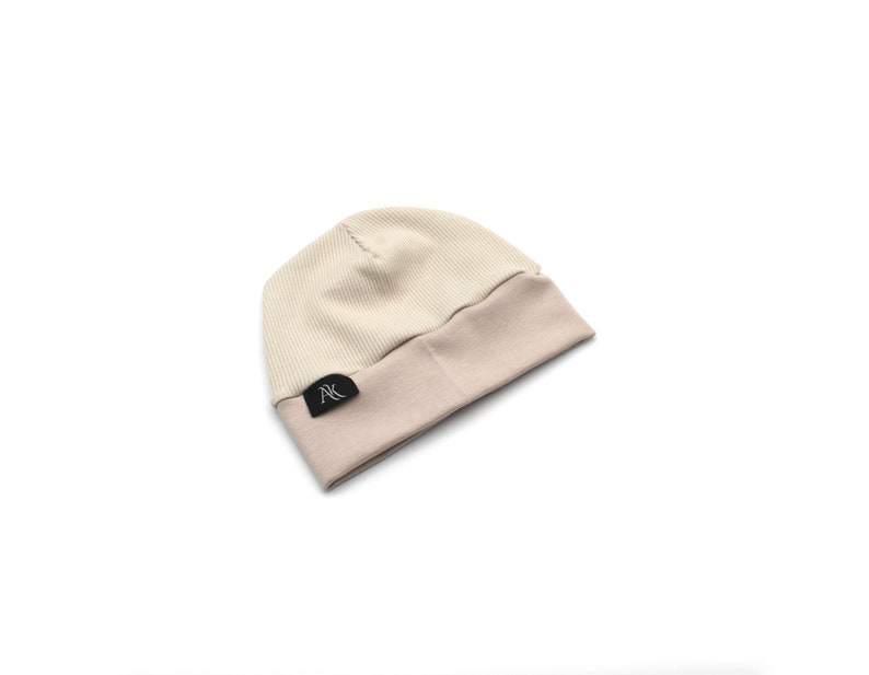 Może przedstawiać: Prążkowana czapka beanie w dwukolorowym designie. G&oacute;rna część jest w kolorze jasnobeżowym, a wywinięty brzeg w ciemniejszym, stonowanym beżu. Mała czarna metka z białym tekstem jest wszyta na brzegu.