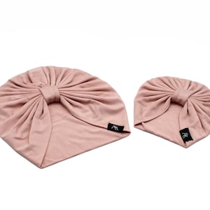May include: Two blush-colored turbans with a gathered knot design. The turbans have a soft, fabric appearance and feature a small black tag with white text. One turban is larger than the other.