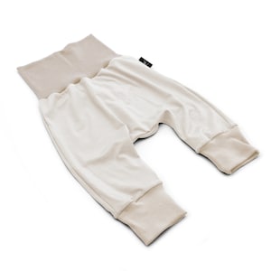 May include: Cream-coloured baby trousers with a wide, tan waistband and matching cuffs. The trousers are made of soft fabric and have a relaxed fit. A small black tag is sewn on the side. Designed for comfort and ease of movement.