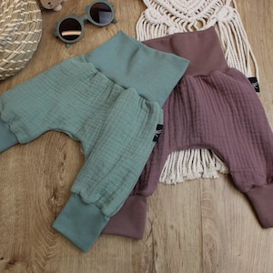 May include: Three pairs of baby pants in shades of teal and mauve. The pants have a high waistband and ribbed cuffs at the ankles. The fabric has a textured, crinkled appearance. The pants are displayed on a light wood surface.
