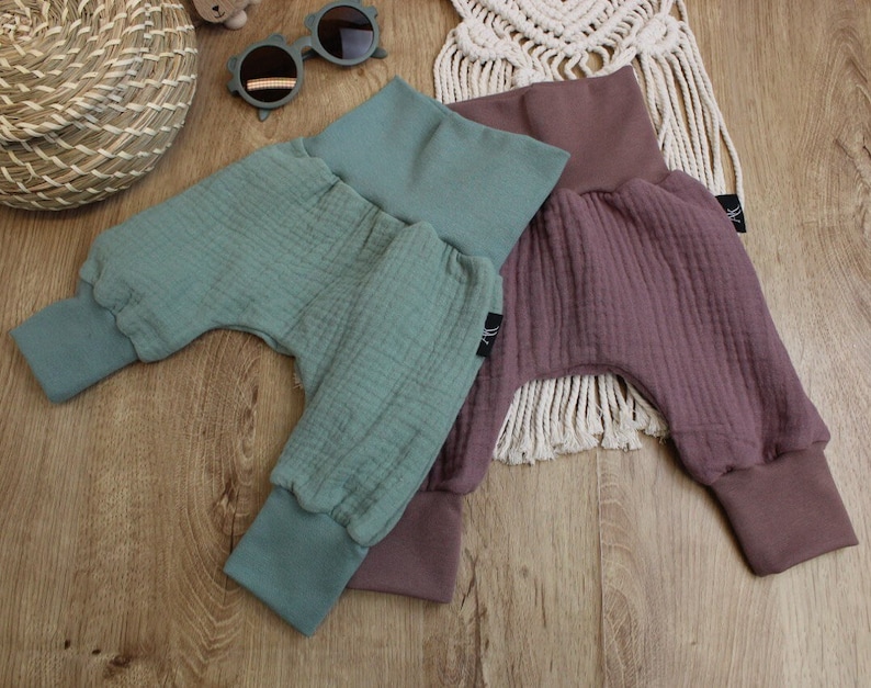 May include: Three pairs of baby pants in shades of teal and mauve. The pants have a high waistband and ribbed cuffs at the ankles. The fabric has a textured, crinkled appearance. The pants are displayed on a light wood surface.