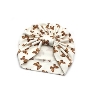 May include: Cream-colored turban with a repeating pattern of brown butterflies. The turban is made of soft fabric and features a knotted bow detail at the front. This stylish headwear is perfect for babies and toddlers.
