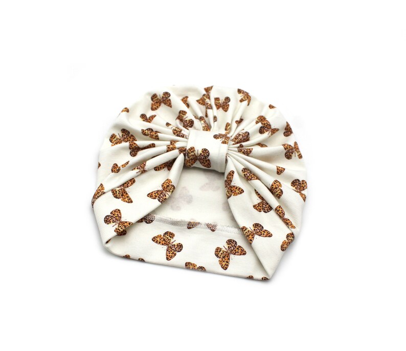 May include: Cream-colored turban with a repeating pattern of brown butterflies. The turban is made of soft fabric and features a knotted bow detail at the front. This stylish headwear is perfect for babies and toddlers.