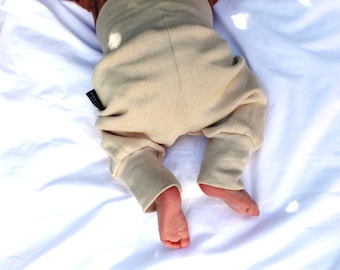May include: Close-up of baby's legs and feet wearing beige knit pants. The pants have a high waist and cuffed ankles. The baby is lying on a white sheet. A small black tag is visible on the pants.