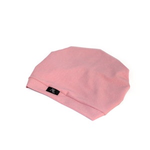 May include: A light pink beanie hat with a soft texture. The hat has a simple design and a small black label with white text. The hat is made of a soft material and is suitable for everyday wear.