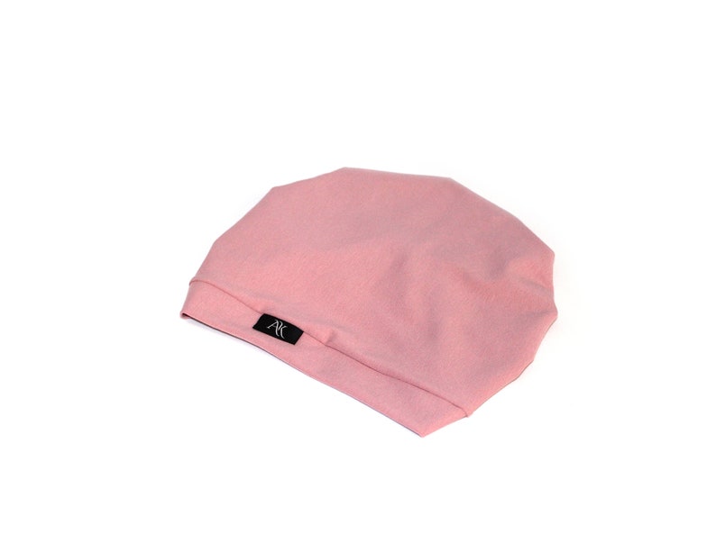 May include: A light pink beanie hat with a soft texture. The hat has a simple design and a small black label with white text. The hat is made of a soft material and is suitable for everyday wear.