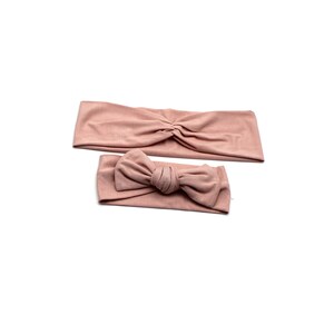 May include: Two blush-colored headbands. One is a wide, knotted style, and the other features a bow. Both are made of soft fabric and are displayed on a white background. These headbands are suitable for various hairstyles.