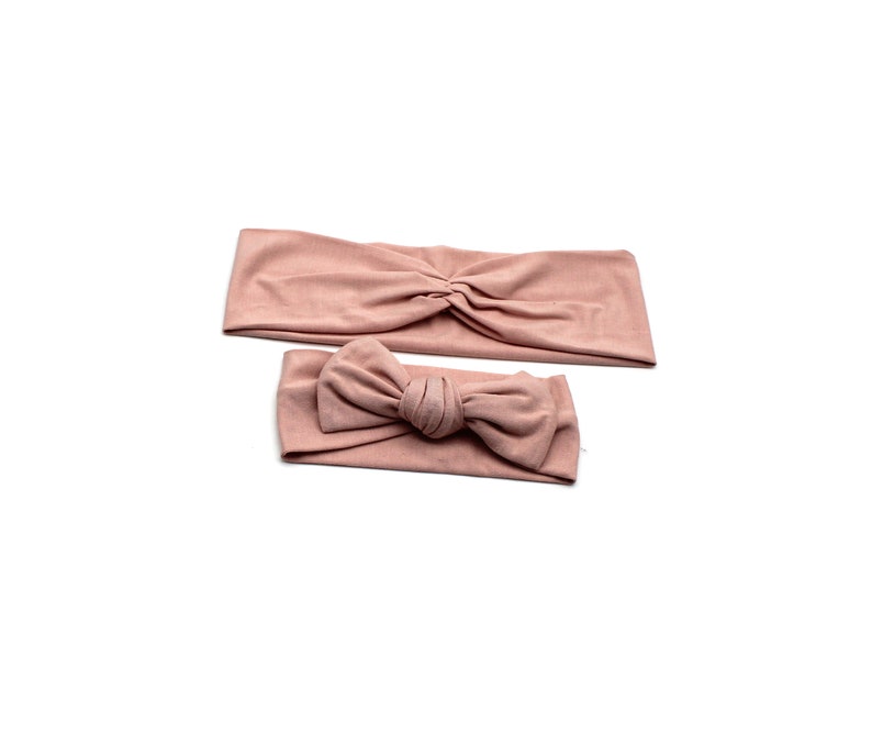 May include: Two blush-colored headbands. One is a wide, knotted style, and the other features a bow. Both are made of soft fabric and are displayed on a white background. These headbands are suitable for various hairstyles.