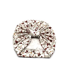 May include: A white turban-style baby hat with a floral pattern. The fabric features small burgundy flowers and green leaves scattered across a cream background. The hat is knotted at the top, creating a stylish accessory.