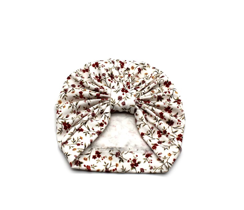 May include: A white turban-style baby hat with a floral pattern. The fabric features small burgundy flowers and green leaves scattered across a cream background. The hat is knotted at the top, creating a stylish accessory.