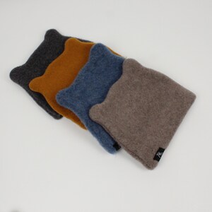 Peut inclure: Quatre chapeaux en laine feutrée dans les tons de gris, jaune moutarde, bleu et taupe sont disposés en diagonale. Chaque chapeau a une forme unique avec un dessus arrondi et une petite extension en forme d'oreille de chaque côté. Une petite étiquette noire est visible sur le chapeau taupe.