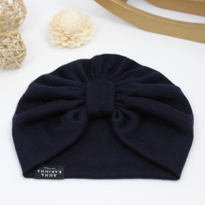 May include: A navy blue turban-style baby hat with a knotted bow detail. The hat has a small rectangular tag with the text "ANNA KARINNA". The hat is made of a soft, ribbed fabric.