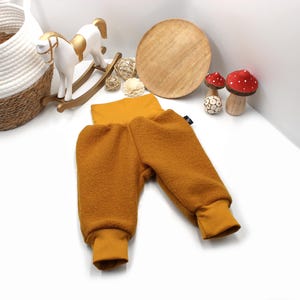 May include: Mustard-coloured baby trousers with a high waistband and ribbed cuffs. The trousers are made of a soft, textured material. The image also shows a white rocking horse, decorative mushrooms, and a wooden circle.
