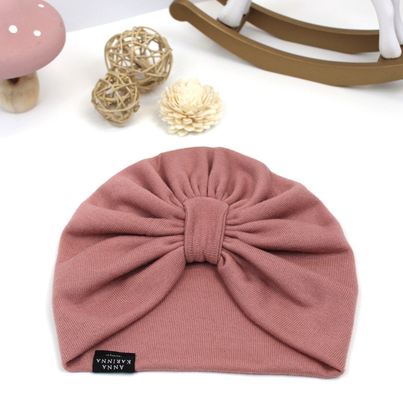 May include: A dusty rose-colored baby turban hat with a knotted bow detail. The hat is made of soft fabric and features a small black label with white text that reads "ANNA KARINNA". The hat is designed for infants and toddlers.