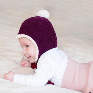 Peut inclure: Un bonnet en tricot violet fonc&eacute; avec une bordure en fausse fourrure blanche le long de l'ouverture du visage et un pompon blanc sur le dessus. Le bonnet poss&egrave;de des liens en tissu violet fonc&eacute; au menton, port&eacute; par un b&eacute;b&eacute; en haut blanc.