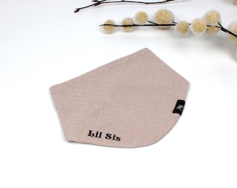 May include: A light tan baby bib with the text "Lil Sis" in black. The bib is made of a soft, textured fabric and has a curved shape. A small black tag with white text is on the right side of the bib.